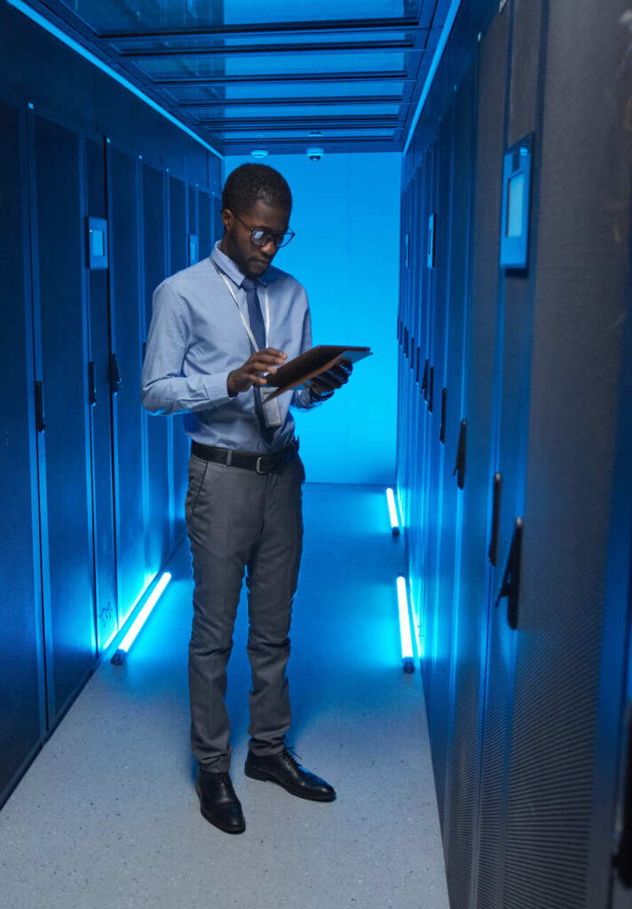 Full length portrait of African American man standing by server cabinet while working with supercomputer in data center and holding tablet, copy space
